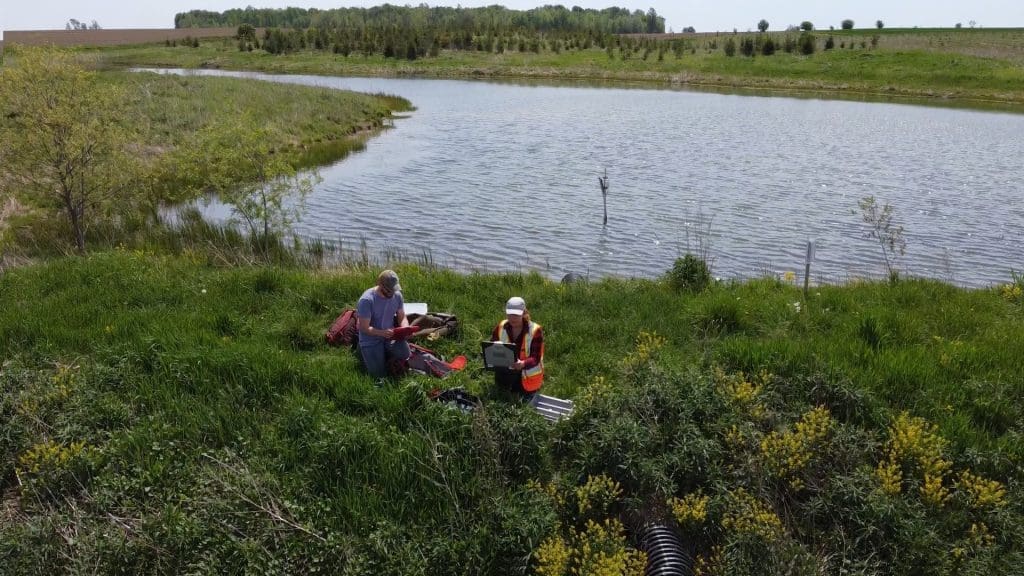 Two scientists kneel beside a small wetland while working with their equipment.