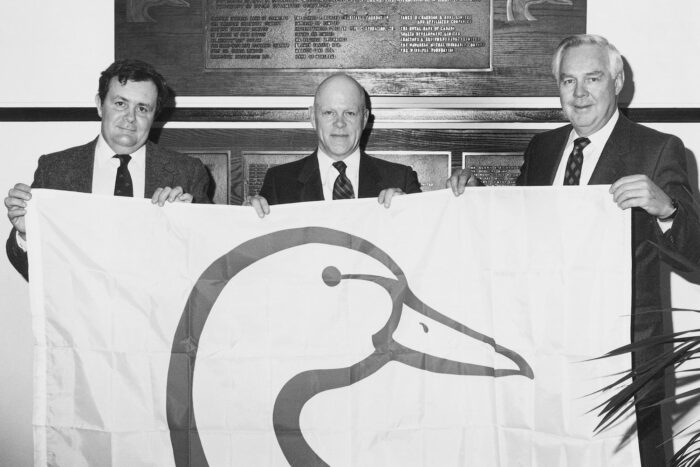 Commercial artist Ernie Wilson (middle), credited with designing the DUC logo, holds a flag alongside former DUC executive vice-president D.S. Morrison (left) and DUC president Jack McKeag (right).