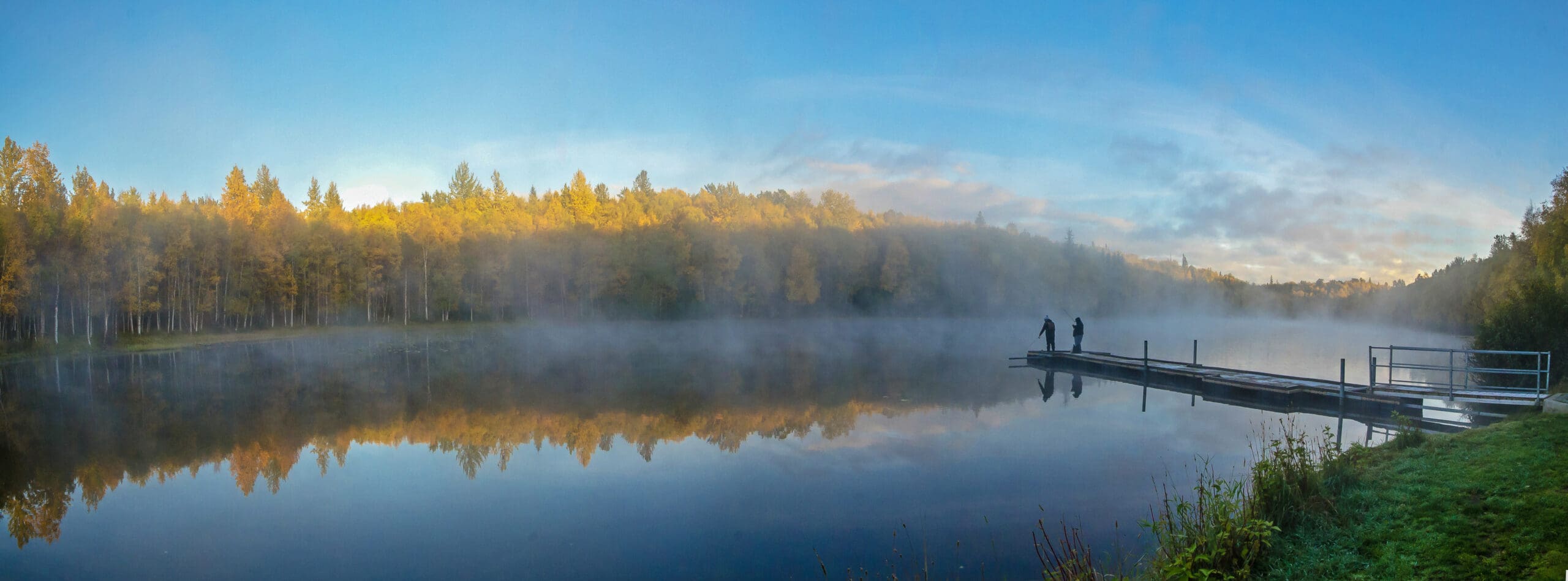 Two people fishing off a dock on a misty lake.