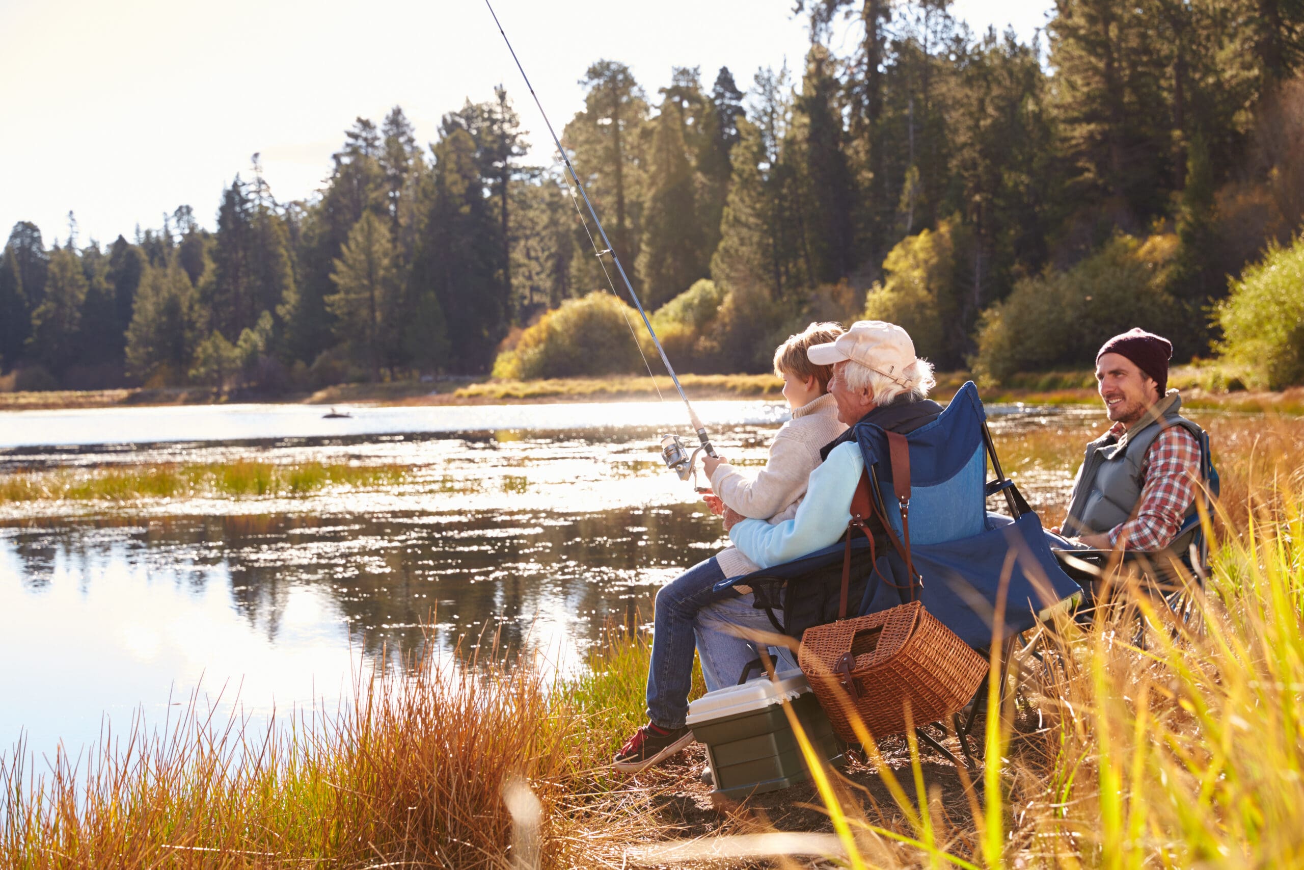 A Grandad teaches his grandson to fish at a lake while dad is watching.